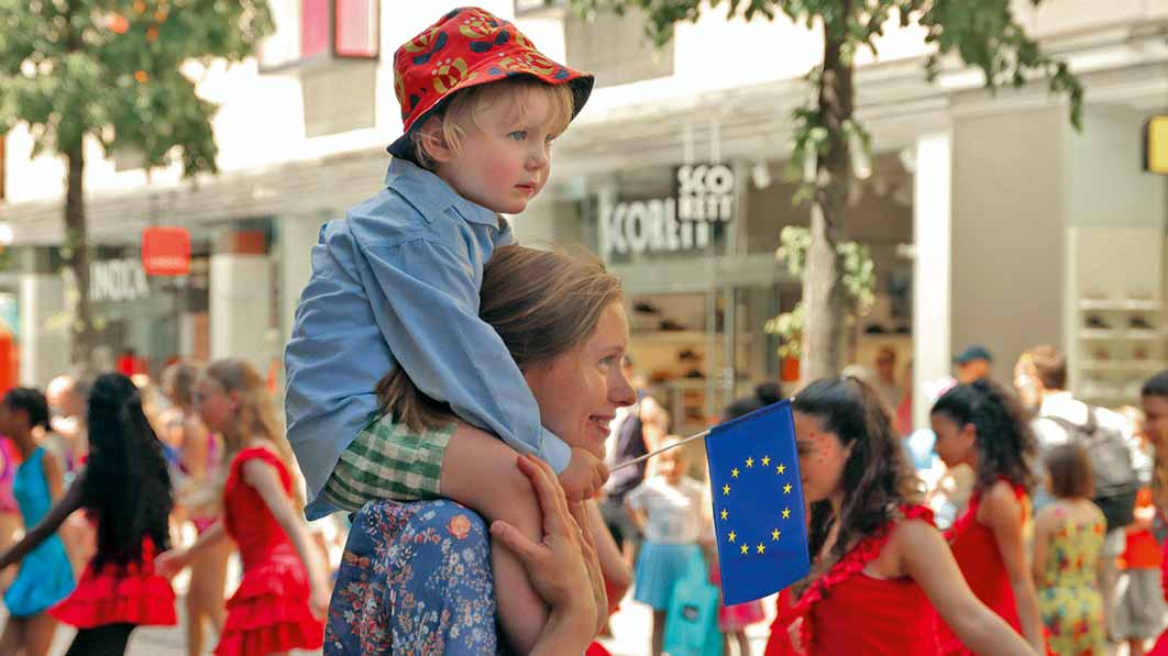 Mutter mit Kind auf den Schultern, das eine EU-Flagge in den Händen hält. 