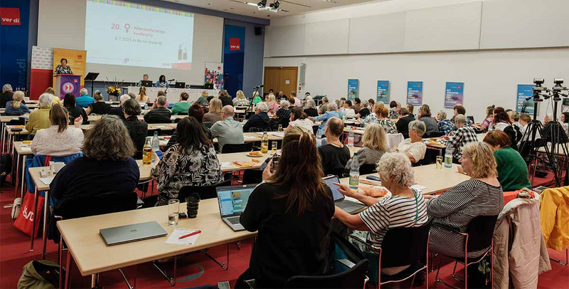 Blick von hinten in einen vollbesetzten Raum. Vorne eine Referenton vor einer Leinwand.
