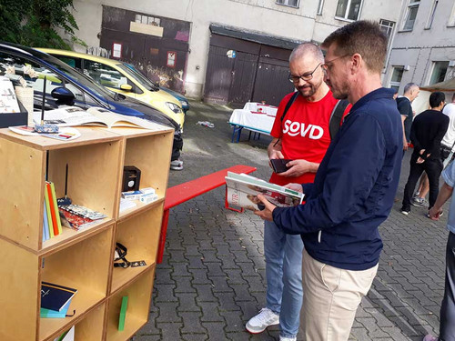 Menschen auf einem Hof vor einem Regal. 