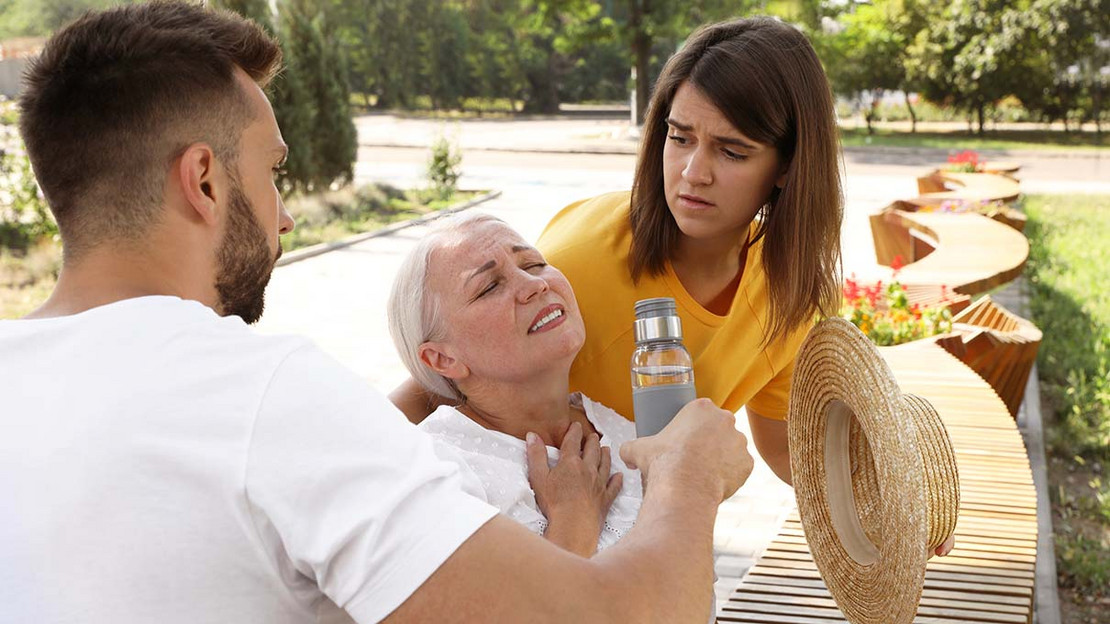Eine alte Frau leidet unter der Hitze. Ein Mann reicht ihr eine Wasserflasche, eine Frau blickt besorgt. 