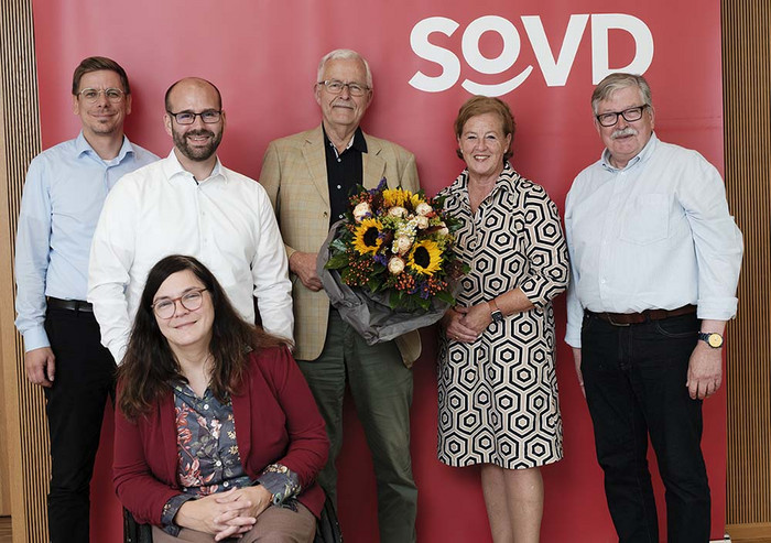 Gruppenbild mit sieben Personen vor einem Aufsteller mit SoVD-Logo. Der Mann in der Mitte hält einen Blumenstrauß in den Händen.