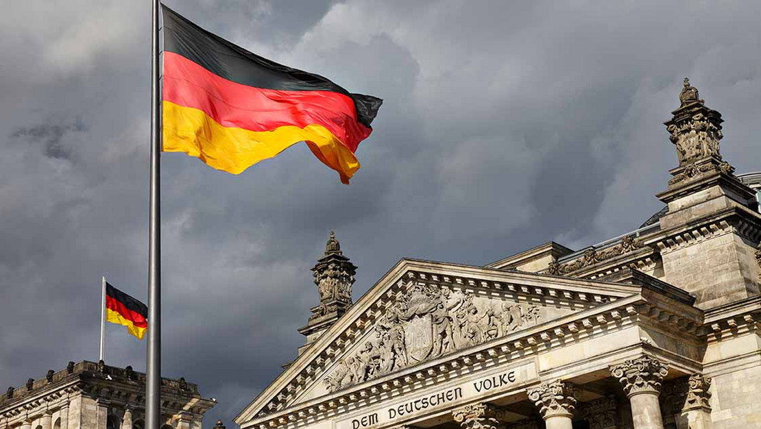 Eingangstor des Reichstags und eine Deutschlandflagge vor einem grauen Himmel.