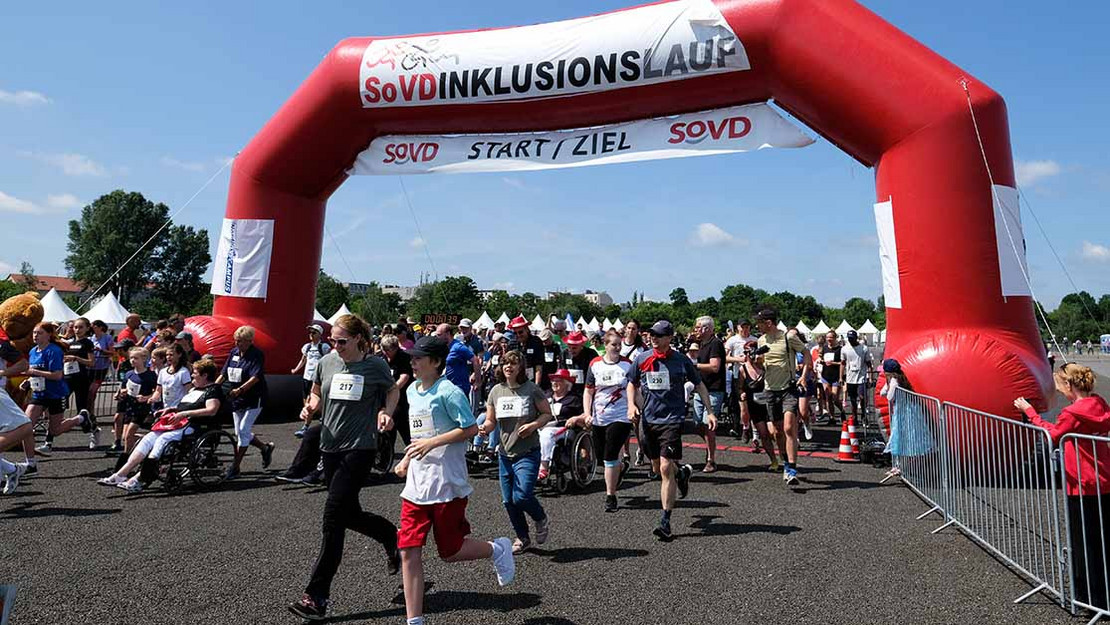 Viele Menschen laufen unter einem Bogen mit der Aufschrift "SoVD-Inklusionslauf: Start / Ziel". 