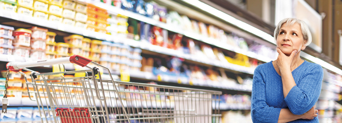 Frau steht neben einem Einkaufswagen vor einem Regal im Supermarkt. 