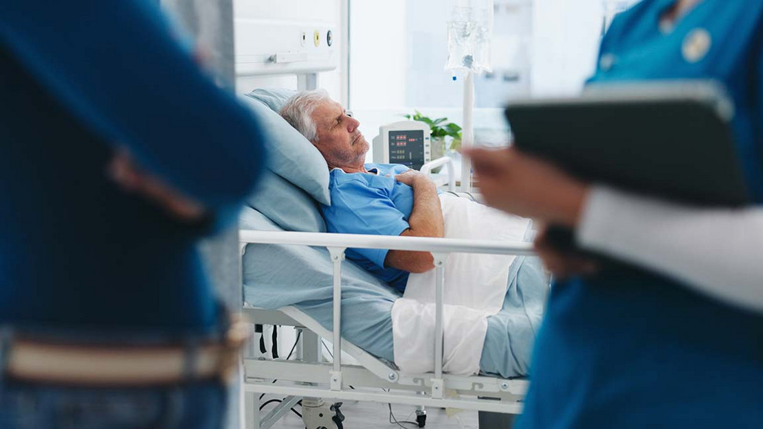 Ein Mann liegt im Krankenhaus und fasst sich an die Brust während im Vordergrund eine Angehörige und eine Medizinerin miteinander sprechen.