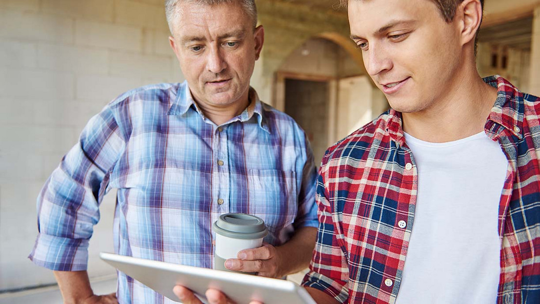 Ein älterer und ein jüngerer Arbeiter blicken auf ein Tablet.