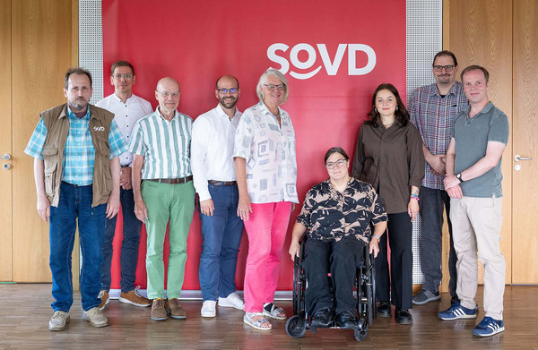 Neun Personen in einem Raum vor einer großen, roten Leinwand mit SoVD-Logo.