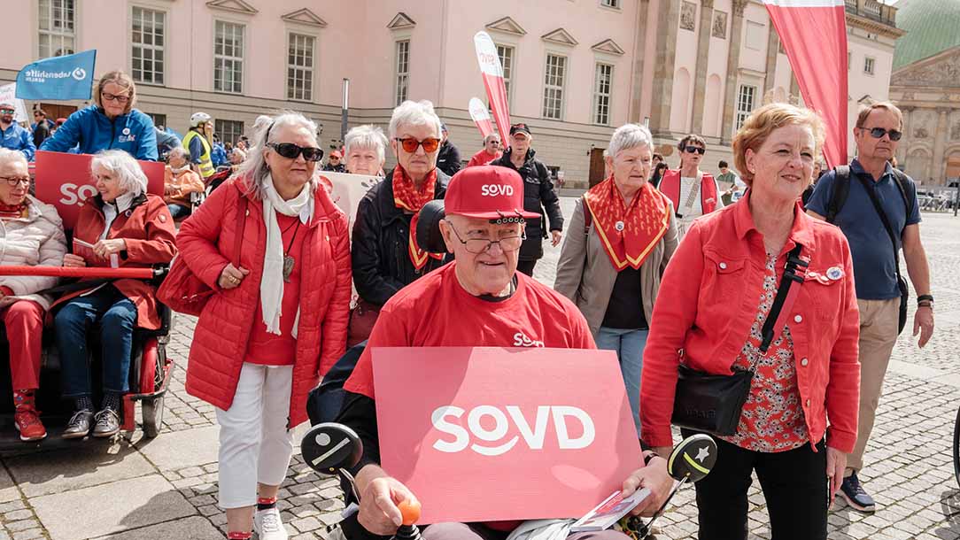 Menschen in roter Kleidung und einem Plakat mit dem SoVD-Logo. 