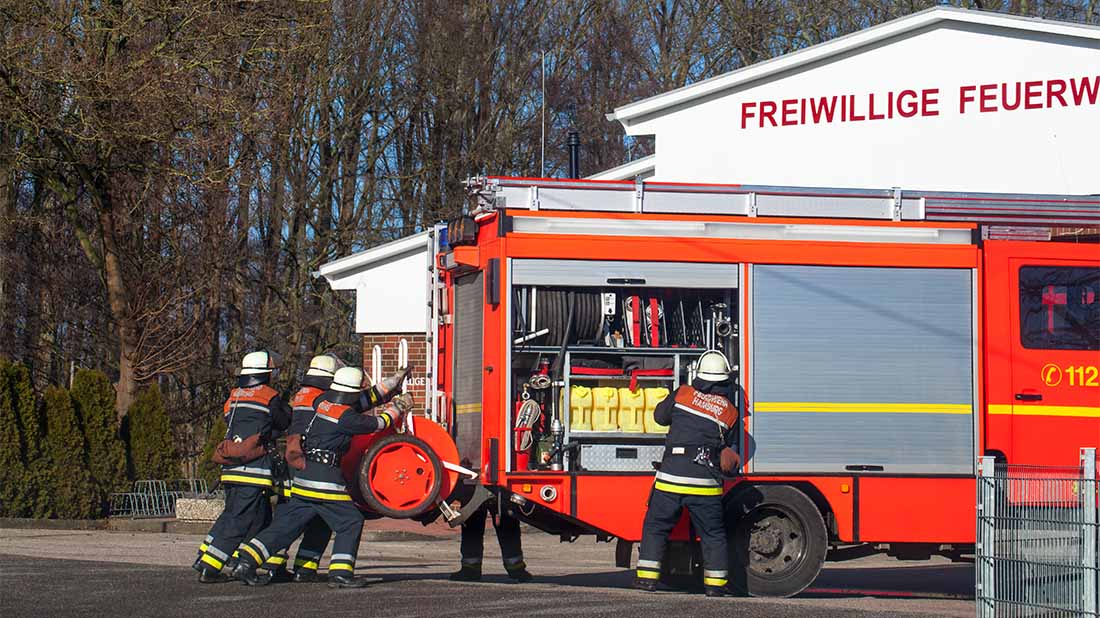 Feuerwehrmänner und ein Löschfahrzezug vor einem Gebäude der Freiwilligen Feuerwehr. 